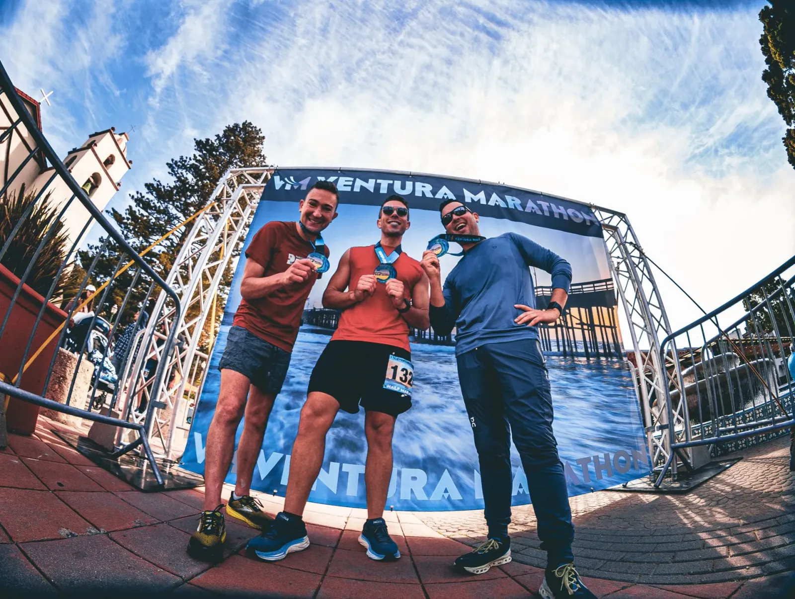 Three smiling runners proudly display their medals in front of the Ventura Marathon banner.