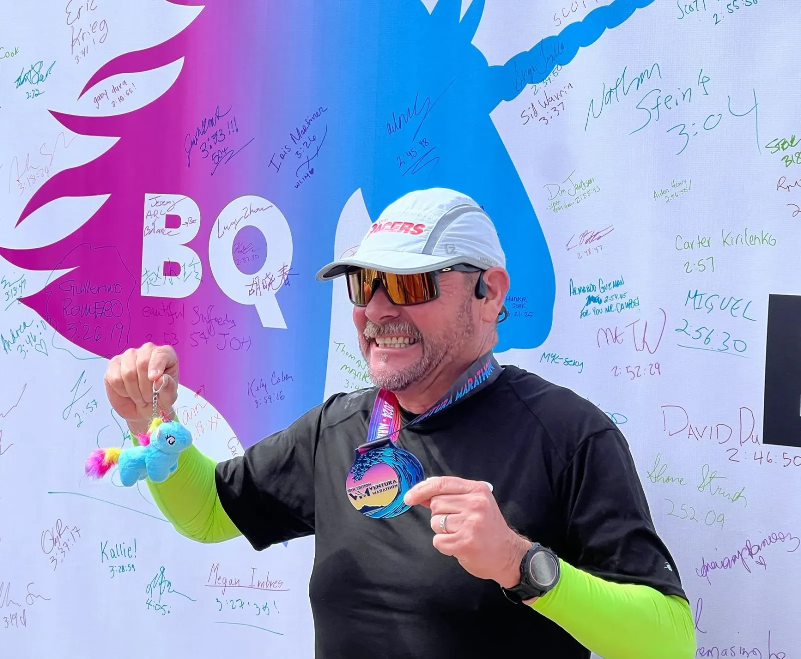 A smiling male runner holds up a medal and a small plush unicorn keychain in front of a wall covered in runner signatures and times.