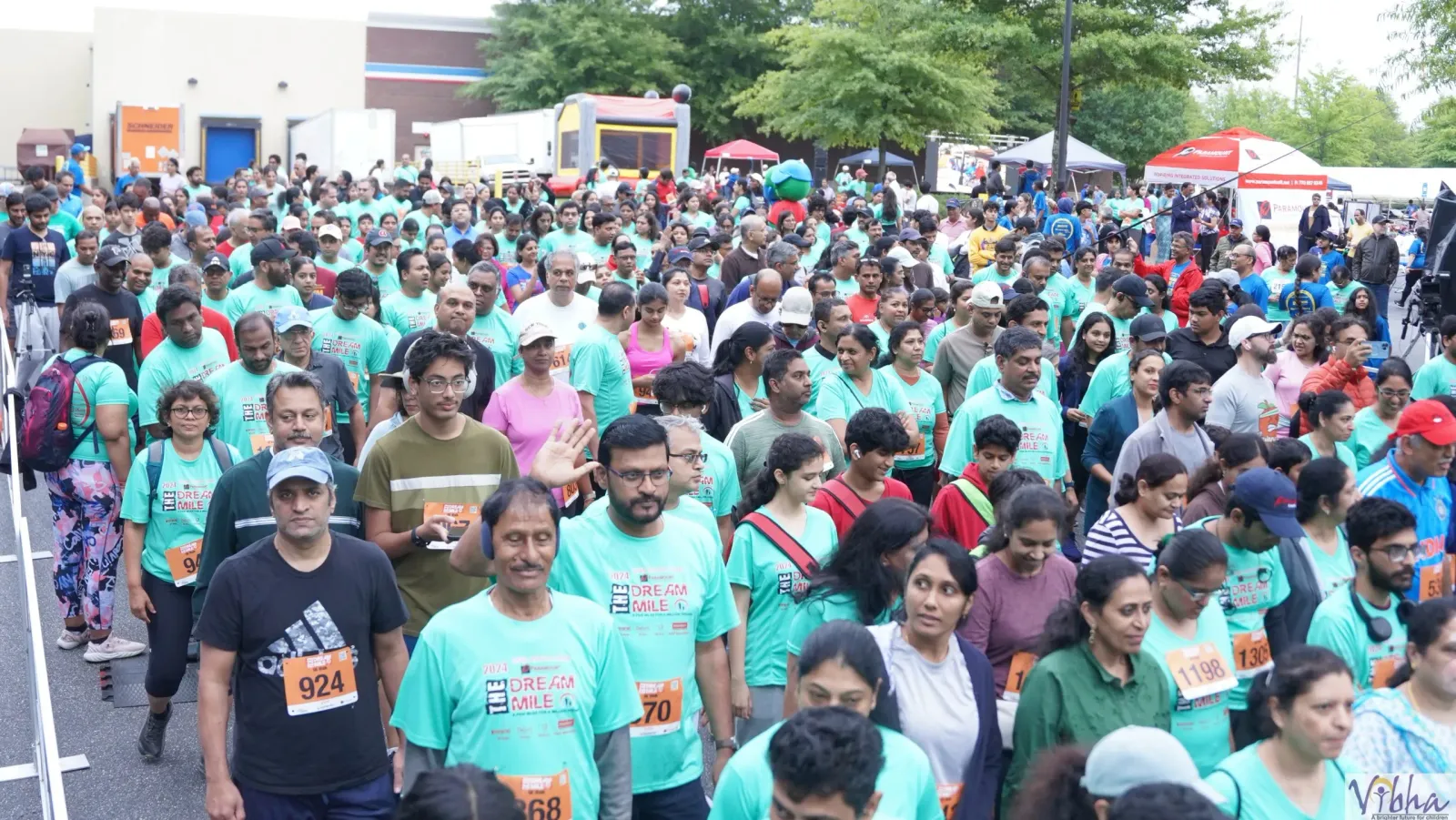 A large group of runners in teal shirts with bib numbers are gathered at the start of the Vibha Dallas Dream Mile race.