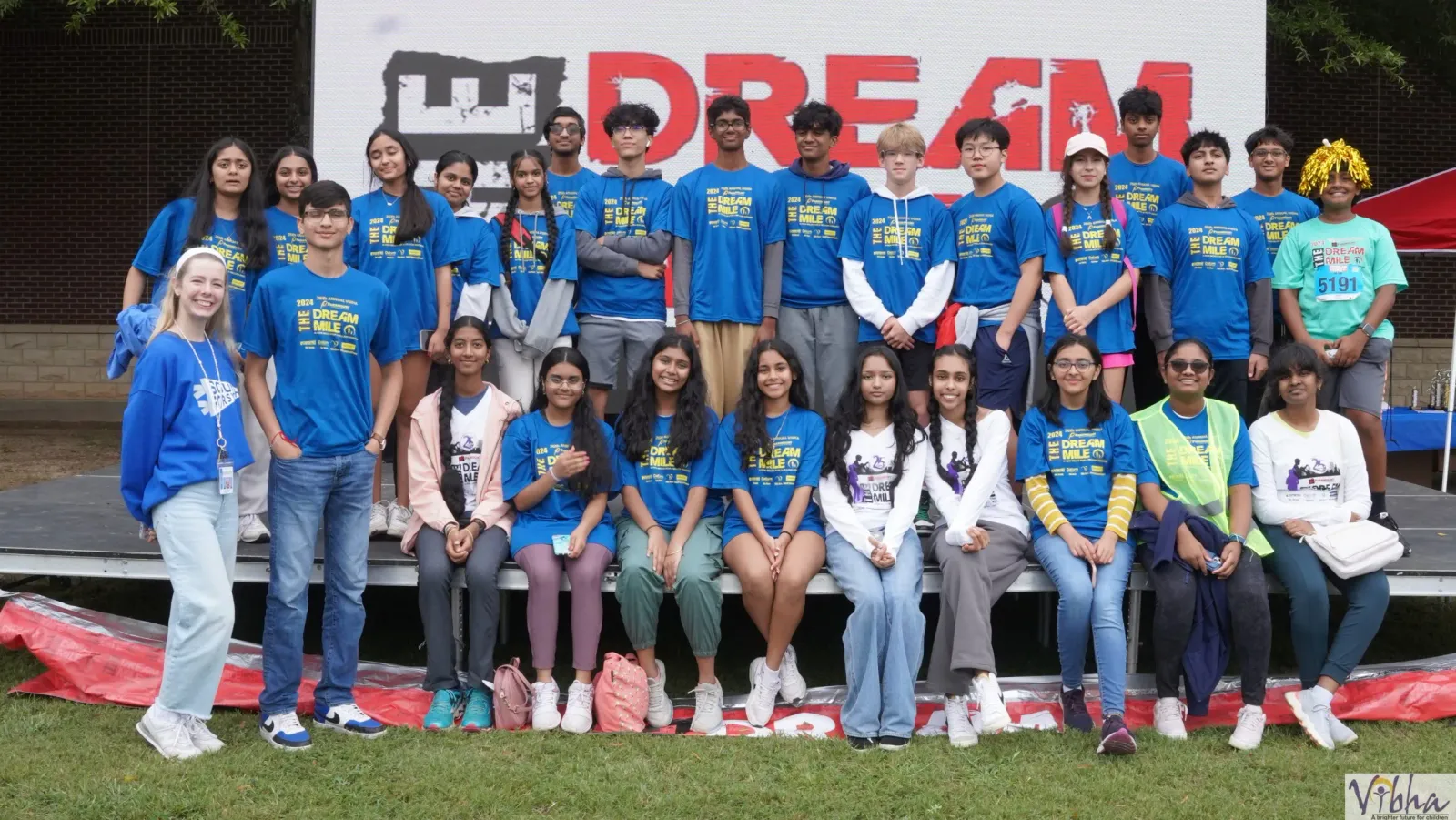 A group of young people in blue t-shirts pose for a photo in front of a large "DREAM MILE" banner.