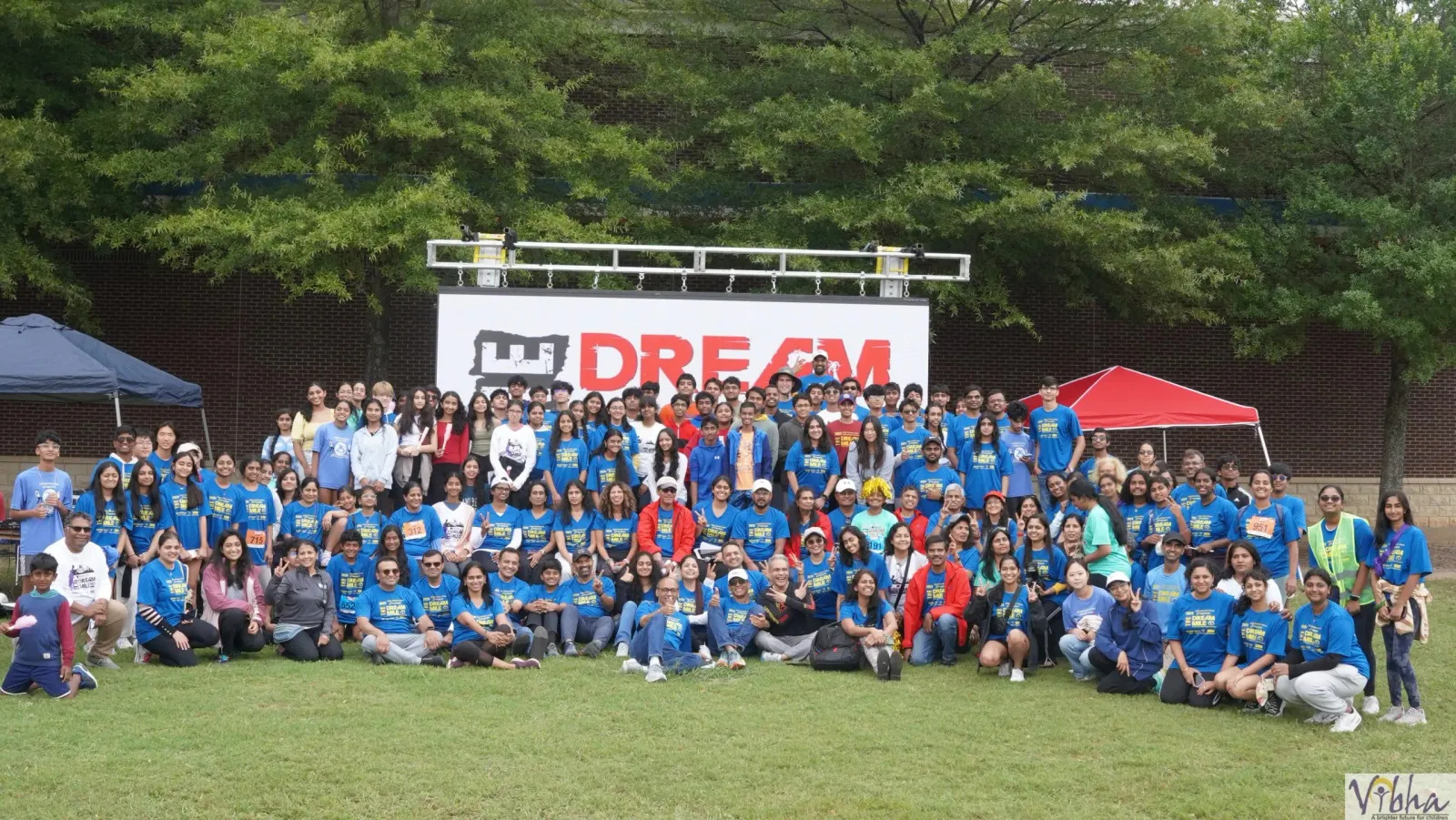 A large group of people, many wearing blue t-shirts with 'DREAM MILE' printed on them, pose for a photo in front of a large 'DREAM' banner.