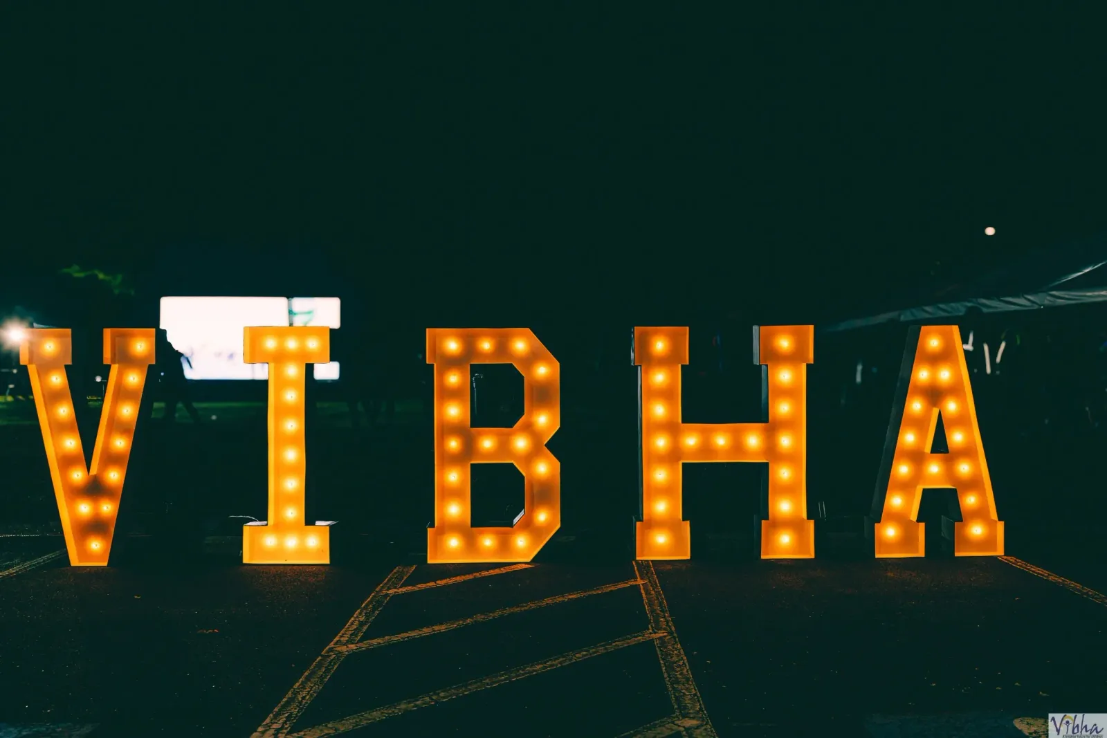 Large illuminated letters spelling 'VIBHA' at night, with blurred figures of people in the background.