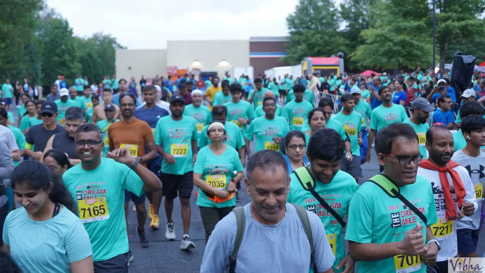 A large group of runners in teal shirts with bib numbers are walking forward at the start of the Dream Mile race.