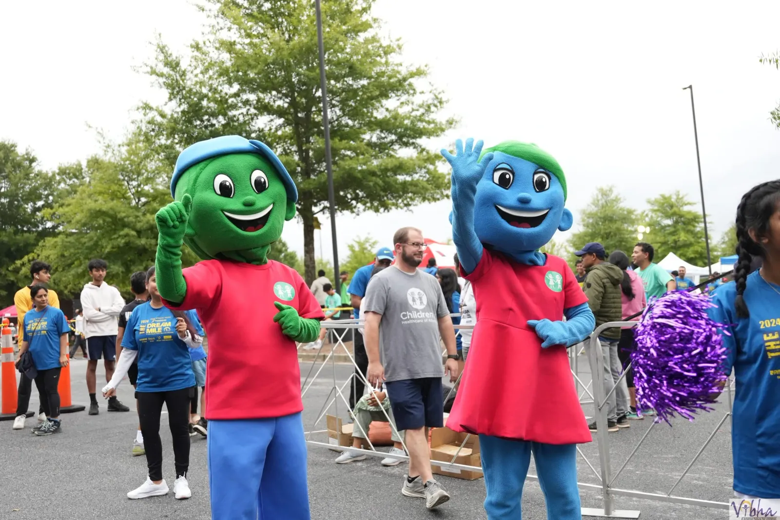 Two race mascots, one green and one blue, wave to the crowd at the Vibha Dallas Dream Mile event. People in race t-shirts are visible in the background.