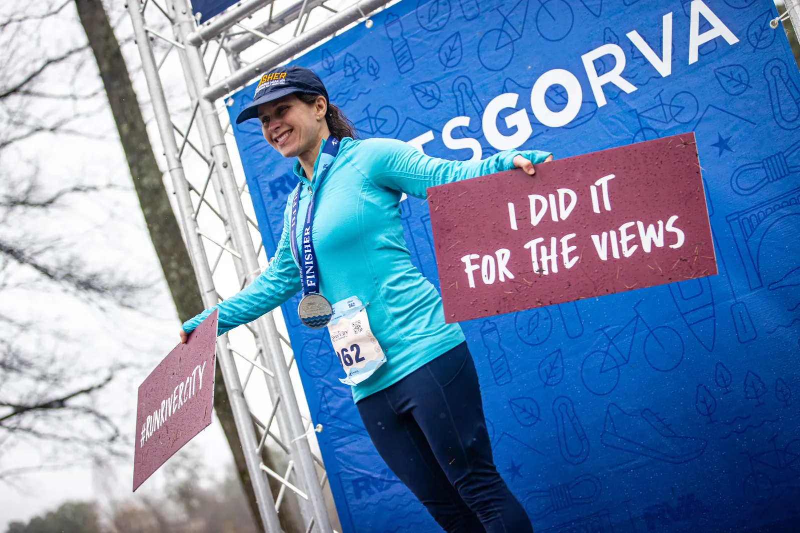 A smiling female runner wearing a blue long-sleeved shirt and a medal around her neck, holding two signs. One sign says "I DID IT FOR THE VIEWS" and the other says "#RUNRIVERCITY". She is standing in front of a blue banner with race graphics.
