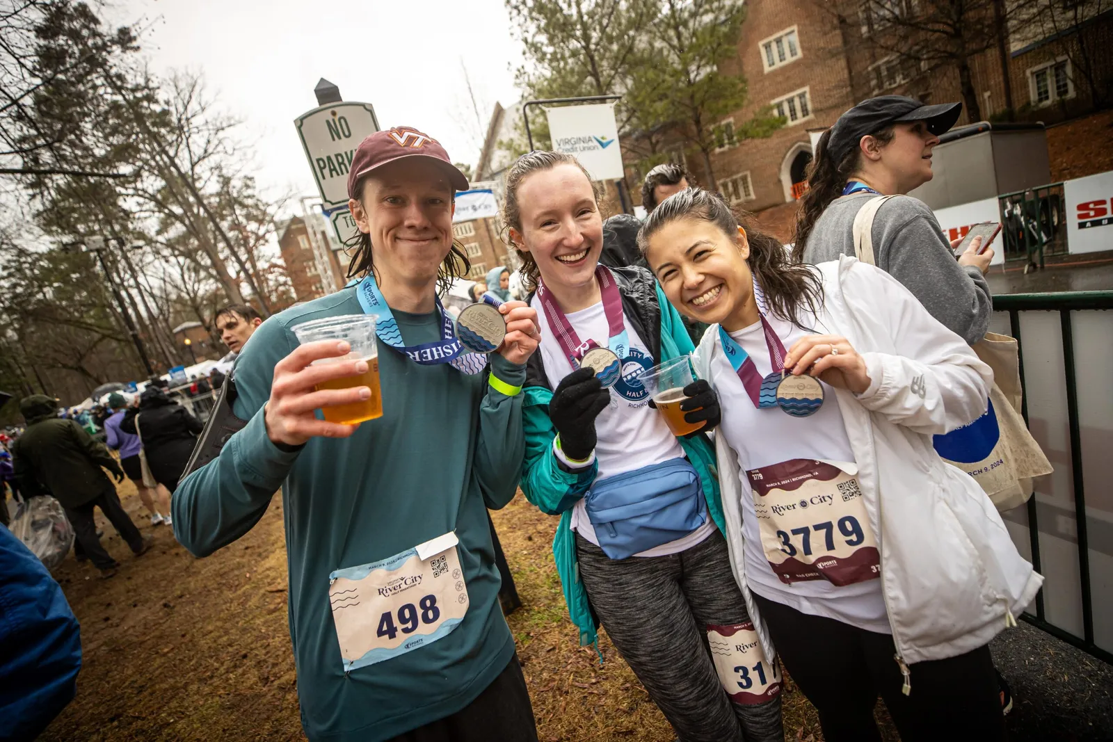 Three smiling runners proudly display their medals and bib numbers after completing the River City Half Marathon, with one holding a drink.
