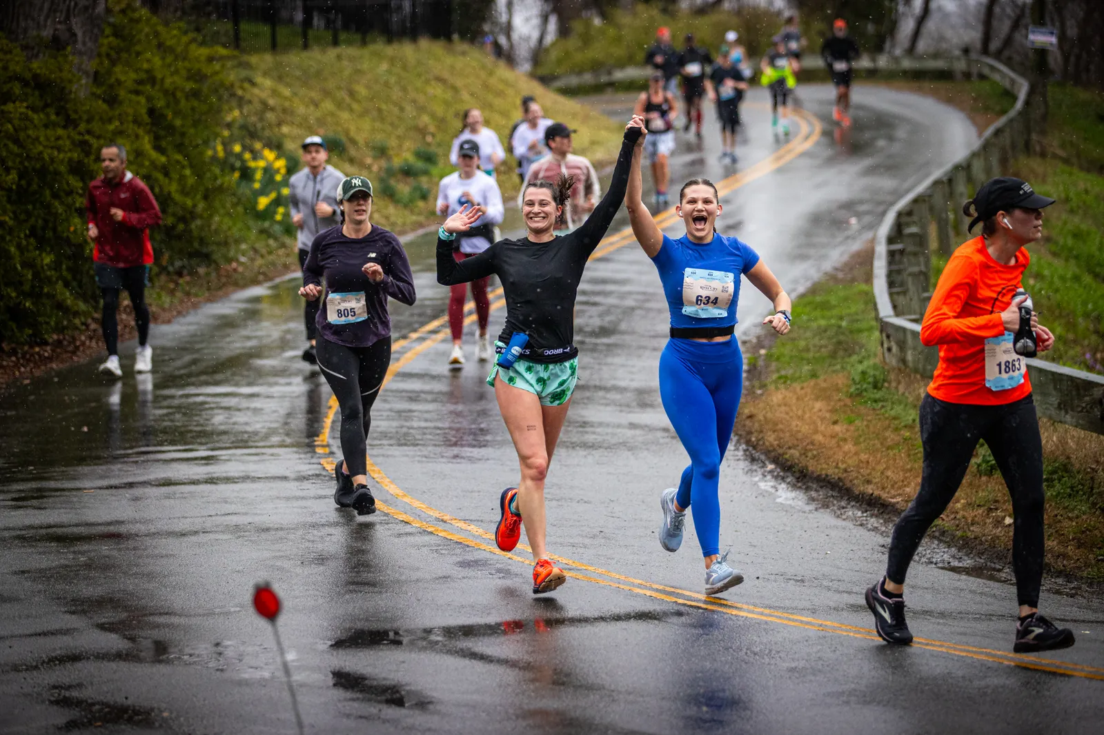 Two female runners celebrate crossing the finish line, holding hands and cheering, with other runners behind them on a wet road.