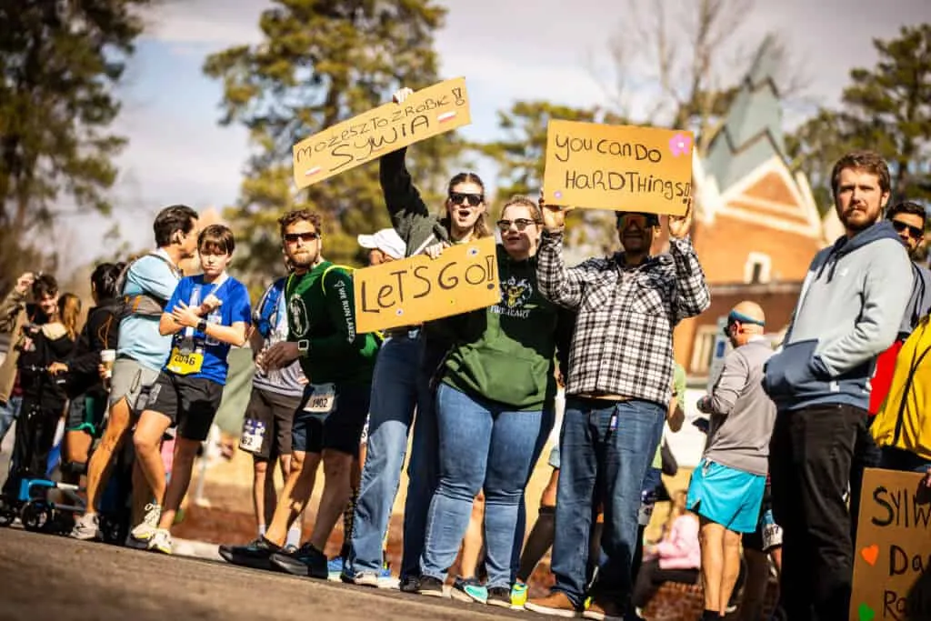 Spectators at a race holding up signs that say "LET'S GO!", "you can do HARD things", and other supportive messages.