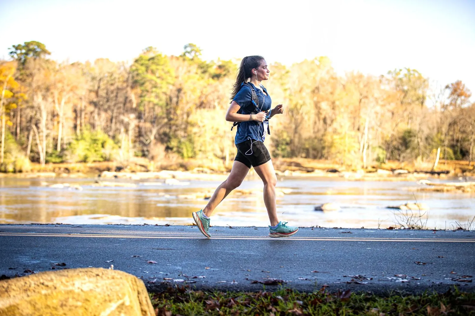 A female runner runs along a road next to a river with trees in the background.