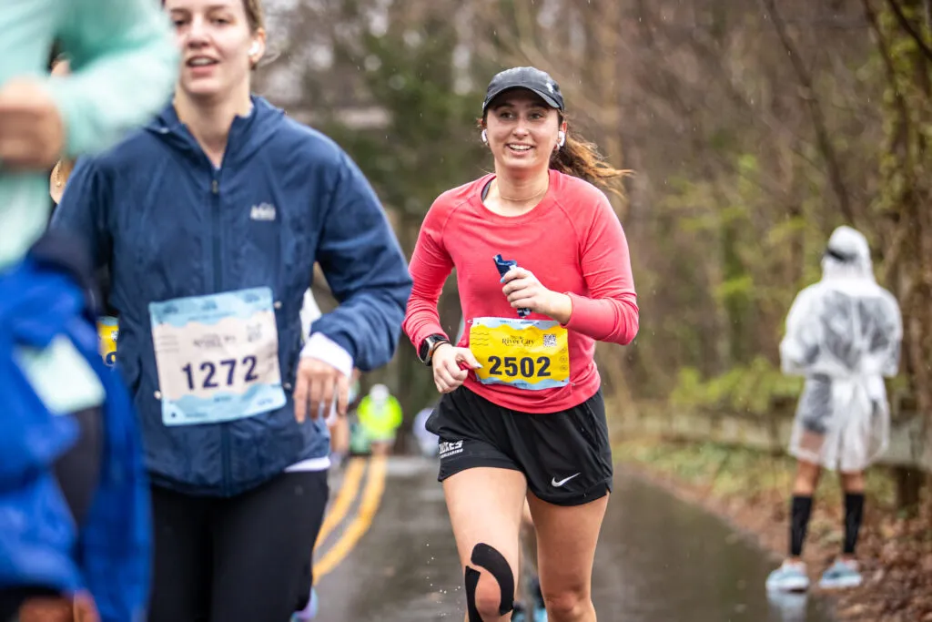A female runner in a pink long-sleeved shirt and black shorts smiles as she runs in a race, with other runners and a person in a rain poncho visible in the background.