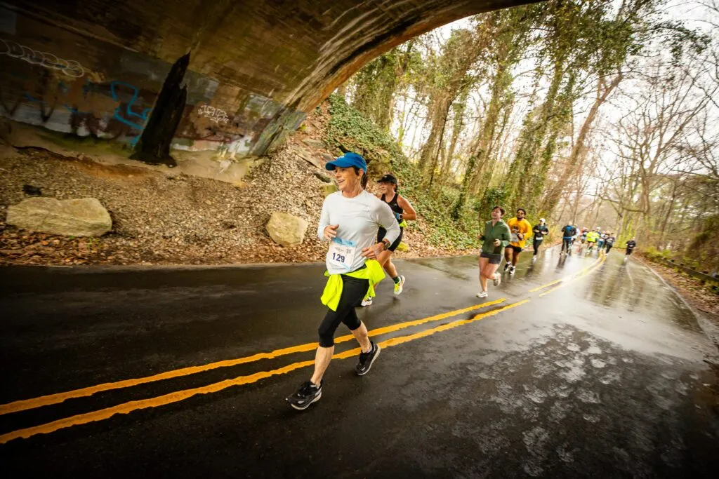Runners participating in the Virginia Credit Union River City Half Marathon and 5K race, running under a bridge on a wet road with trees lining the course.