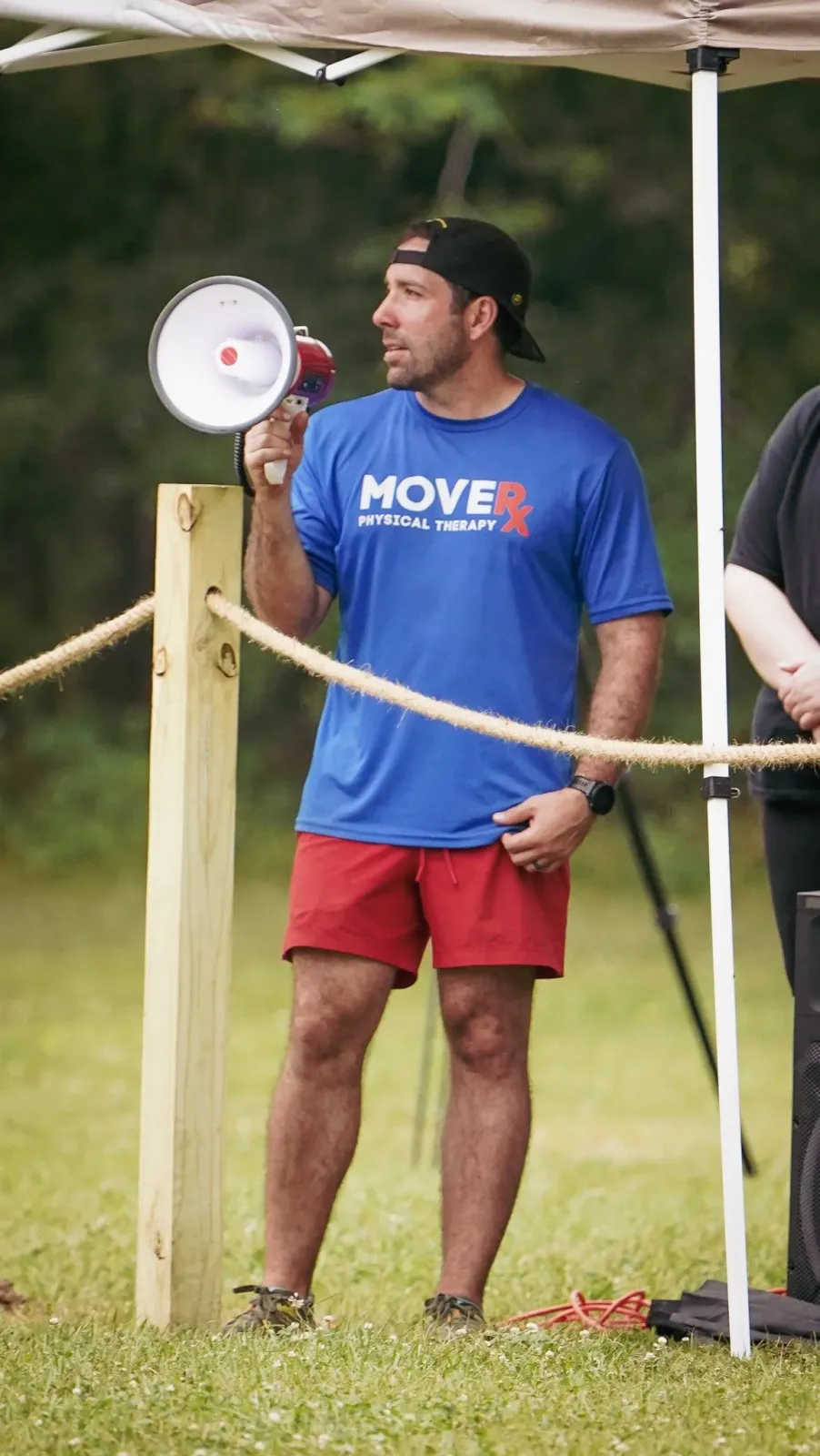 A man in a blue t-shirt and red shorts holds a megaphone, standing near a rope barrier at an outdoor event.
