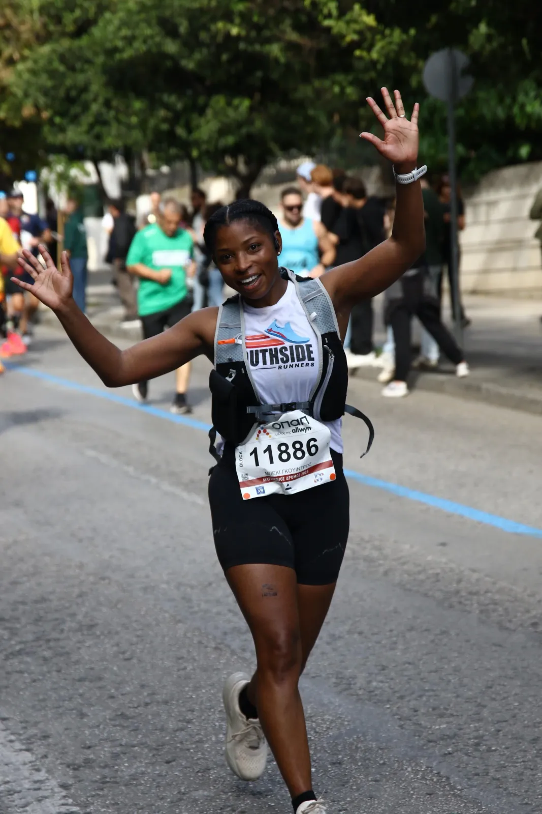 A female runner with a big smile and her hands raised in a wave, running on a road during a race.
