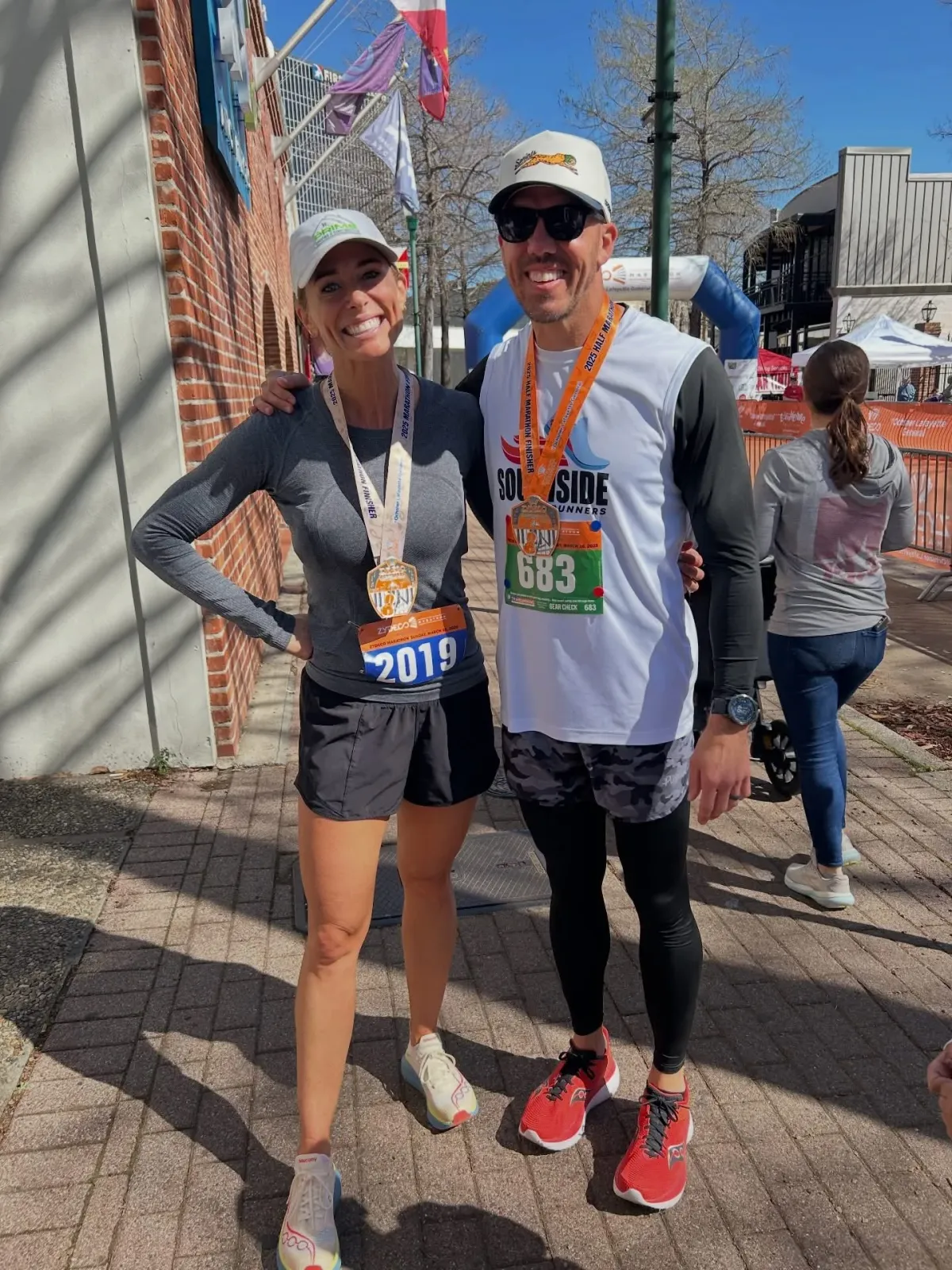 A smiling couple, both wearing race medals and bibs, pose for a photo after completing a half marathon.