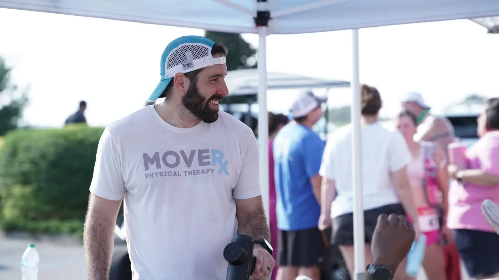 A man wearing a white t-shirt with 'MOVER PHYSICAL THERAPY' and a blue and white cap smiles while looking to the side, holding a massage gun, with other people in the background at an outdoor event.