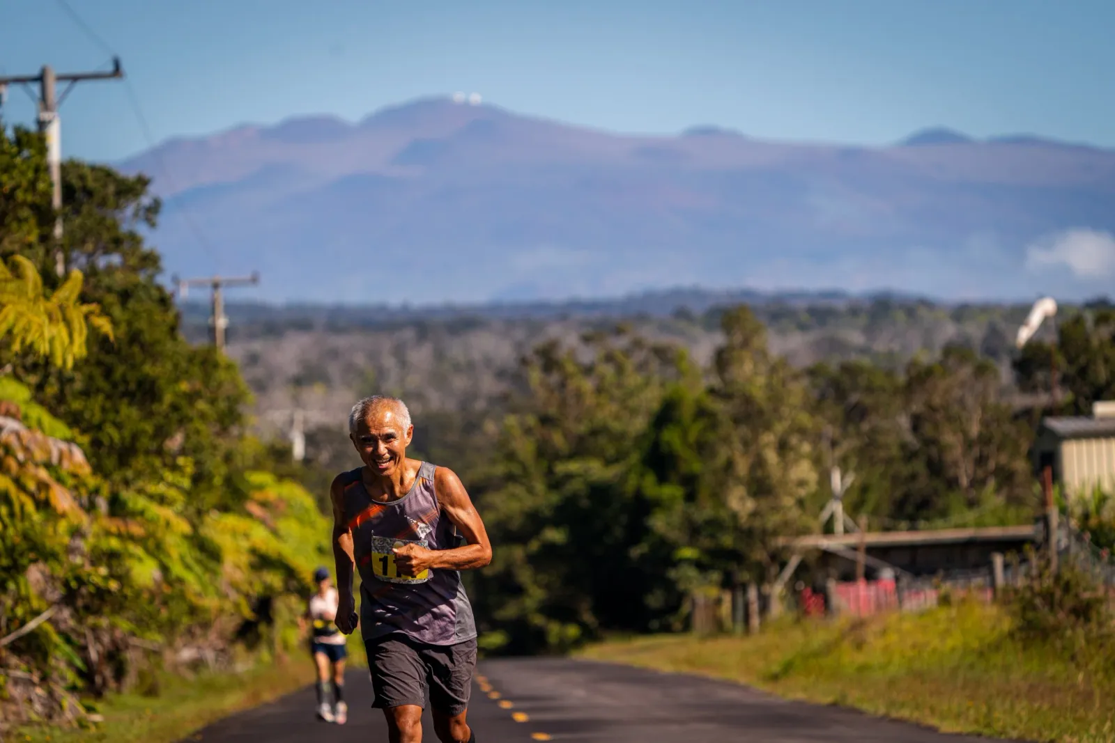 A smiling male runner with bib number 11 runs on a road with a scenic mountain landscape in the background.