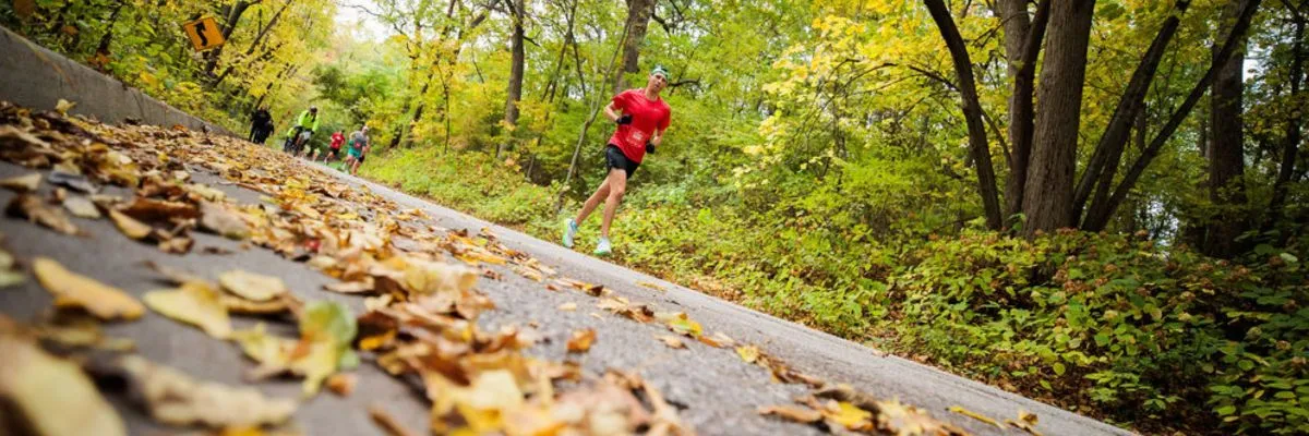 A runner in a red shirt runs on a road covered in fallen autumn leaves, with trees and foliage in the background.
