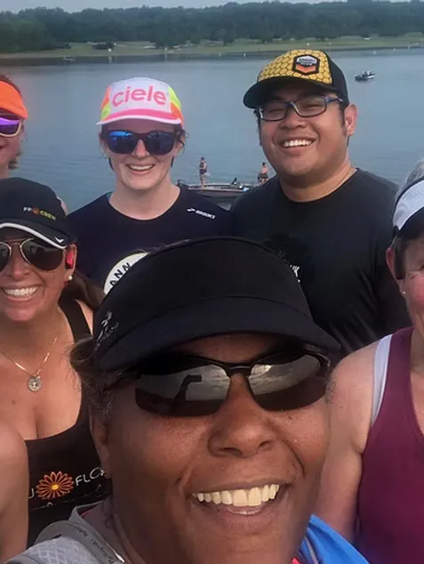 A group of smiling runners pose for a photo with a body of water and trees in the background.