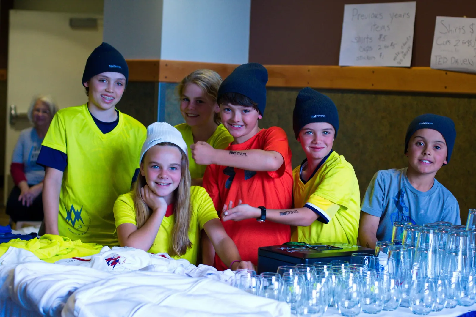 A group of smiling children wearing beanies and race shirts, some with temporary tattoos, stand behind a table with drinks and merchandise.