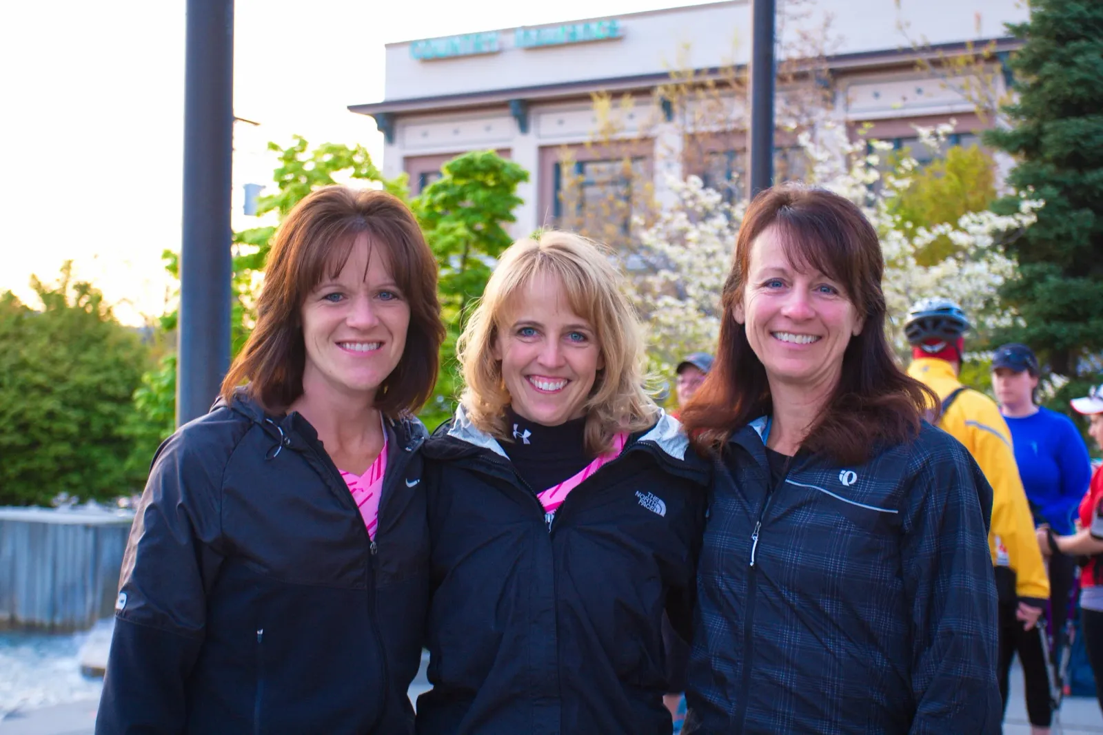 Three women smiling at the camera, wearing athletic jackets, with other runners in the background.