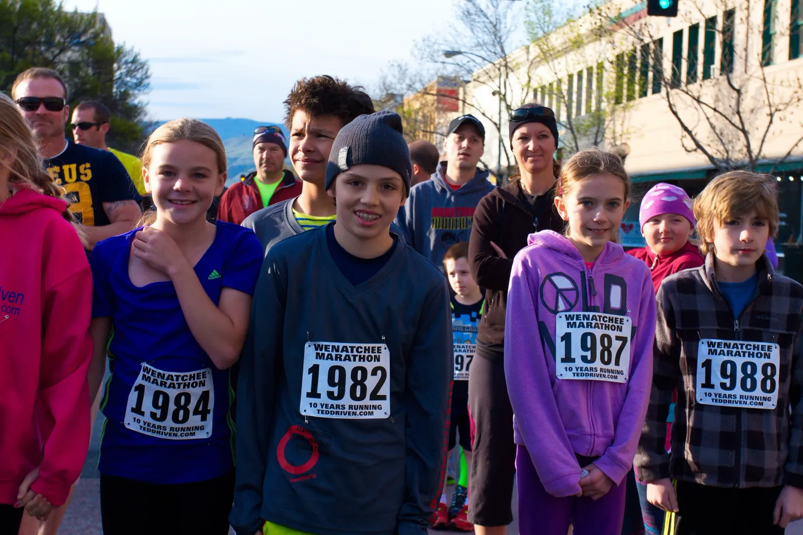 A group of young runners and spectators, some wearing bib numbers, stand together at the start of the Wenatchee Marathon.