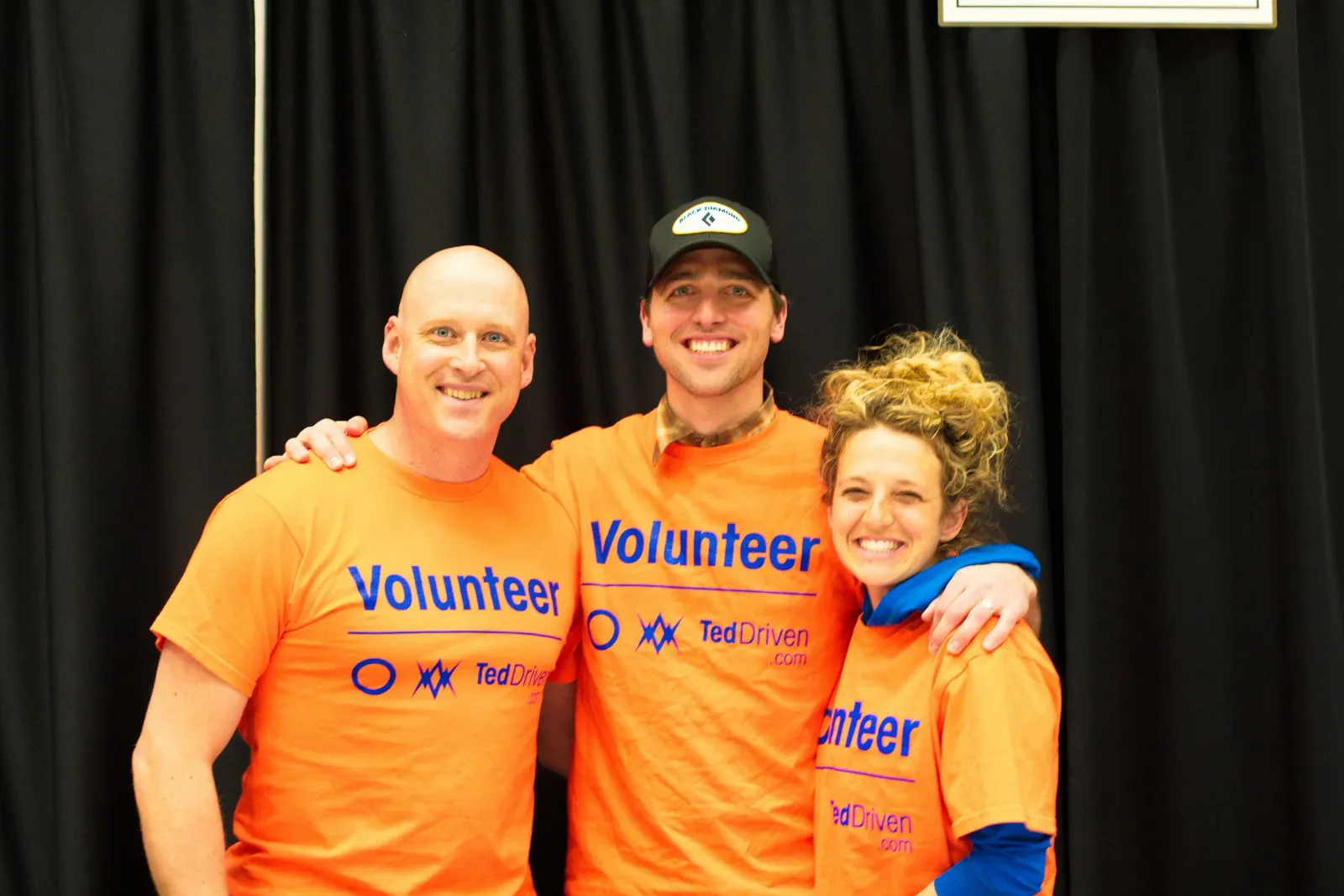 Three smiling volunteers wearing orange 'Volunteer' shirts stand together.