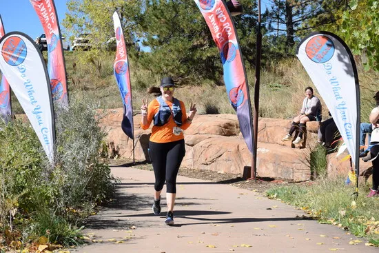 A female runner in an orange shirt and black leggings runs on a paved path, smiling and holding up two peace signs. Race banners with the text 'Do What You Can' are visible on either side of the path.