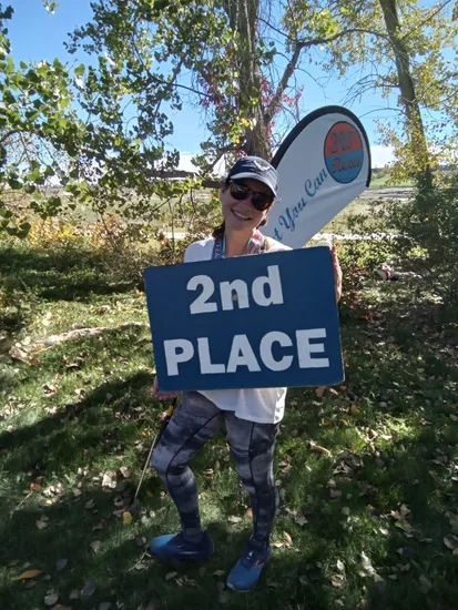 A smiling woman wearing running attire and sunglasses holds a blue sign that says '2nd PLACE' and a race banner is visible behind her.