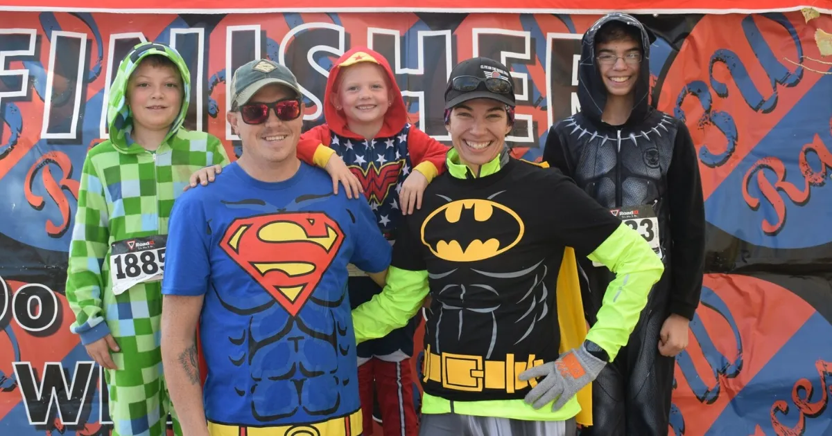 A group of five people, including children and adults, dressed in superhero costumes (Superman, Wonder Woman, Batman, Black Panther) and a Minecraft costume, posing for a photo in front of a race banner.