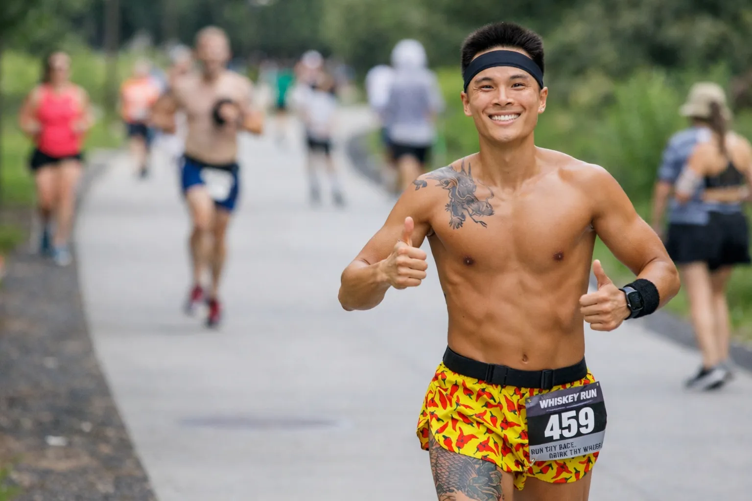 A shirtless runner with a bandana and colorful shorts smiles and gives two thumbs up while running a race.