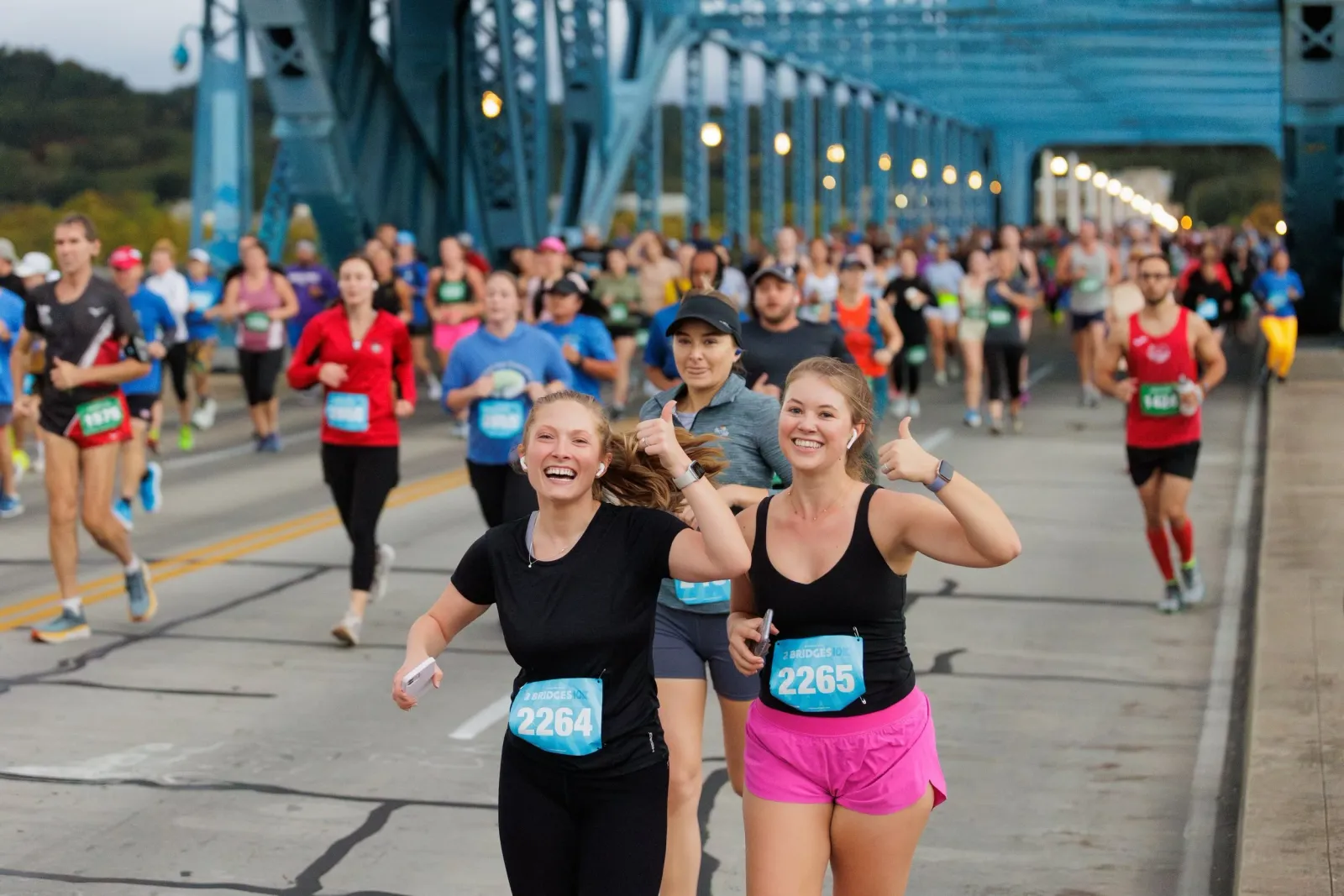 Two female runners smiling and giving thumbs up while running across a bridge during a race, with a crowd of other runners in the background.