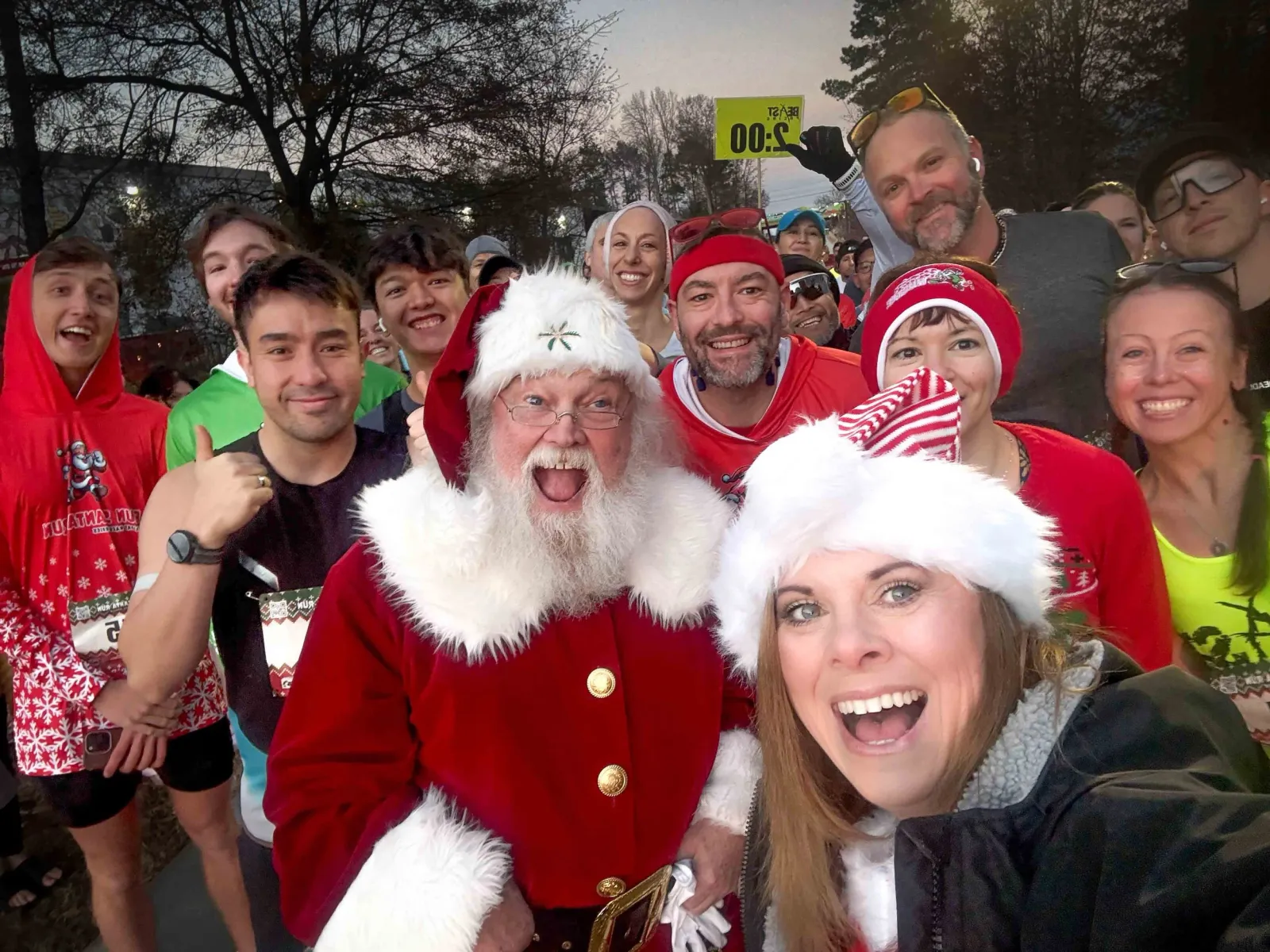 A group of runners, including one dressed as Santa Claus, smiling and taking a selfie at the start line of a festive race.