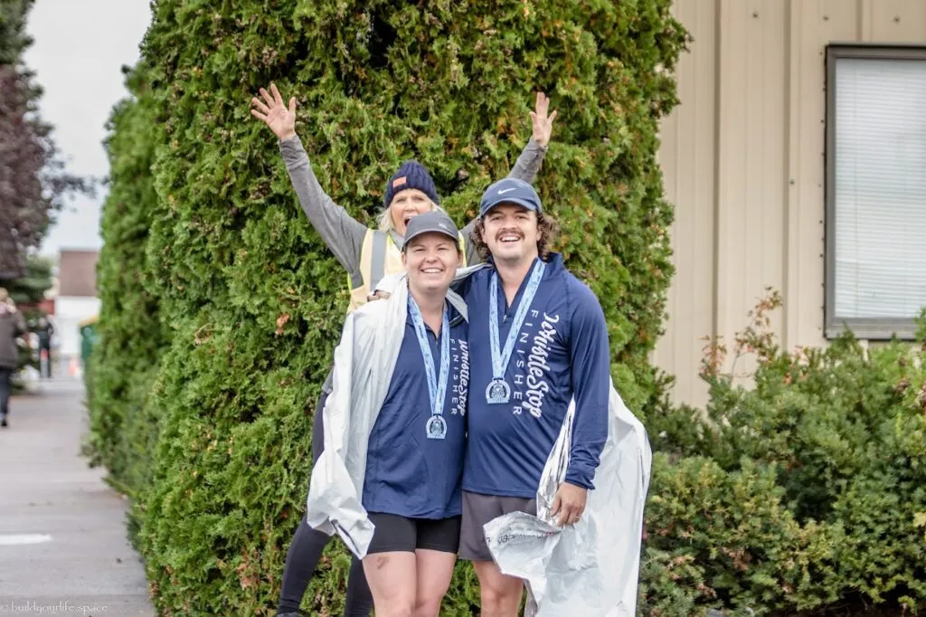 Two smiling runners, a man and a woman, wearing Whistle Stop Finisher medals and dark blue long-sleeved shirts, pose for a photo with a woman in the background waving.