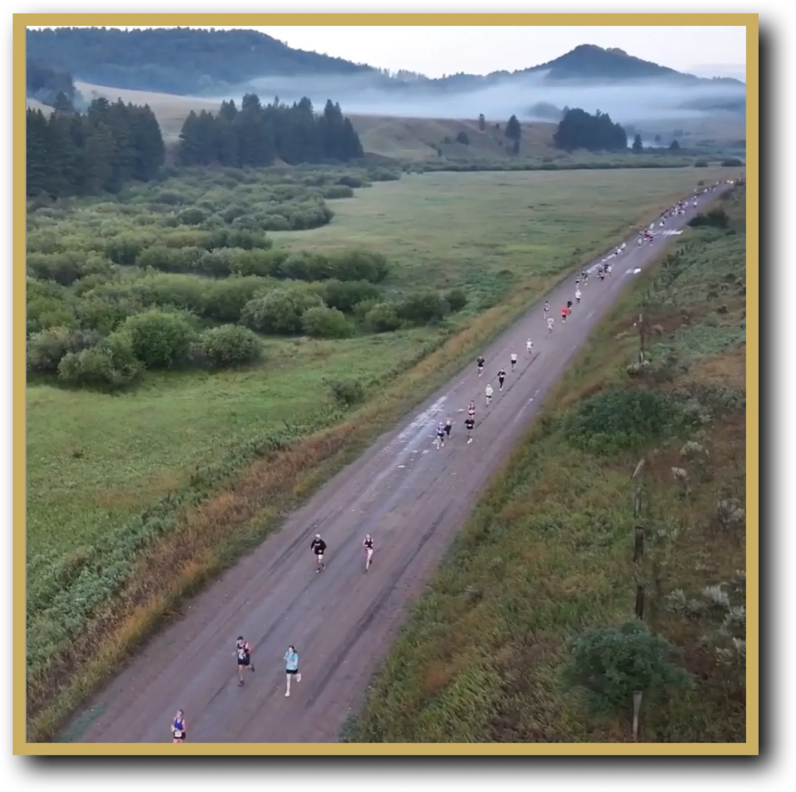 Runners on a dirt road with misty mountains and green fields in the background.