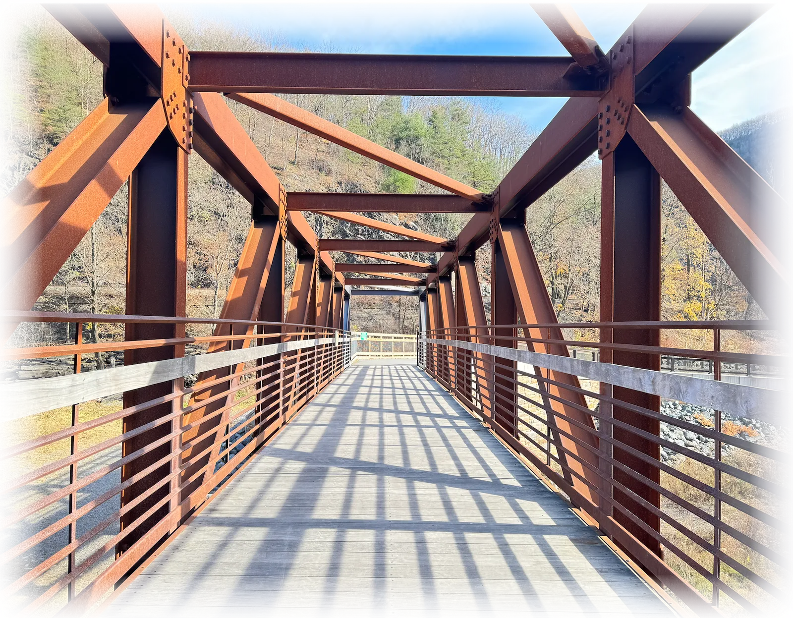 A rusty metal truss bridge with a wooden walkway, casting long shadows on a sunny day.