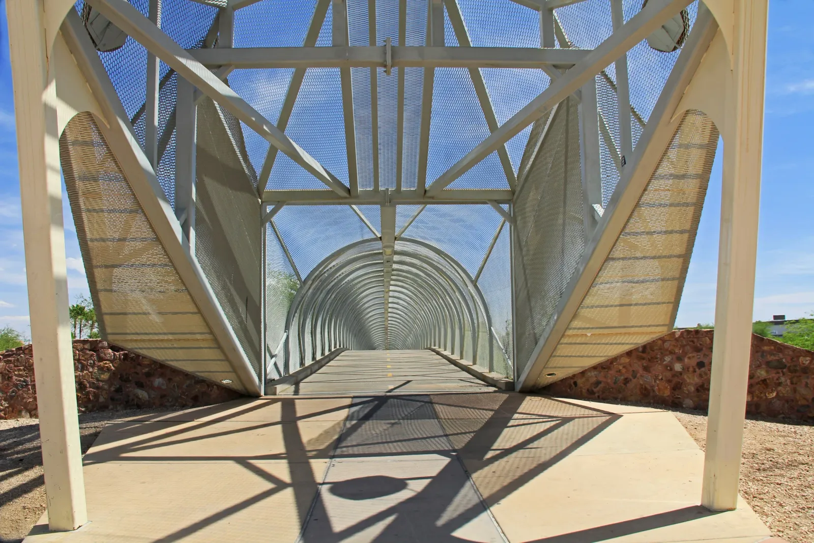 A modern pedestrian bridge with a tunnel-like structure and metal mesh sides, leading into the distance under a clear blue sky.