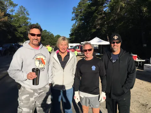 Four people smiling at the camera, with trees and tents in the background, suggesting a race event.