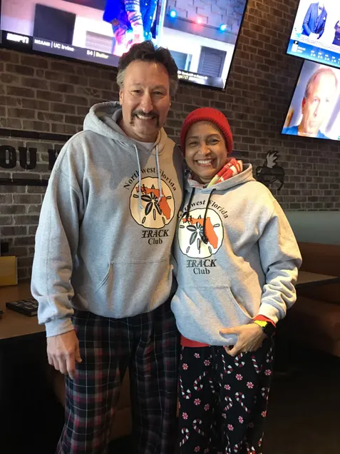 A man and a woman smiling at the camera, both wearing grey hoodies with a 'Northwest Florida Track Club' logo.