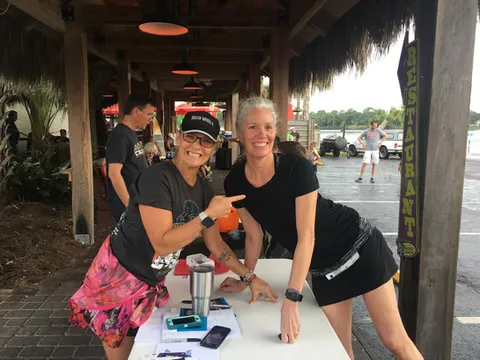 Two smiling women at a race event, one pointing towards the other.