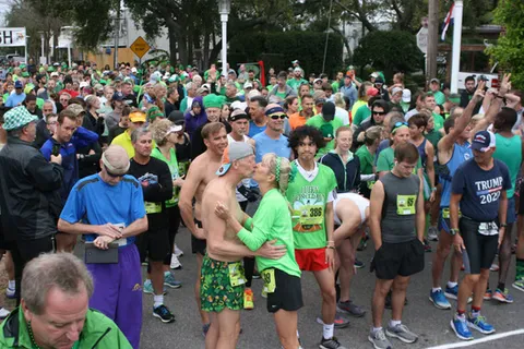 A large crowd of runners in green attire gathered at the start line of a race, with a couple kissing in the foreground.