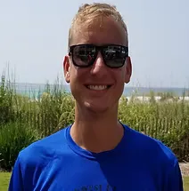 A young man wearing sunglasses and a blue t-shirt smiles at the camera with a beach and ocean in the background.