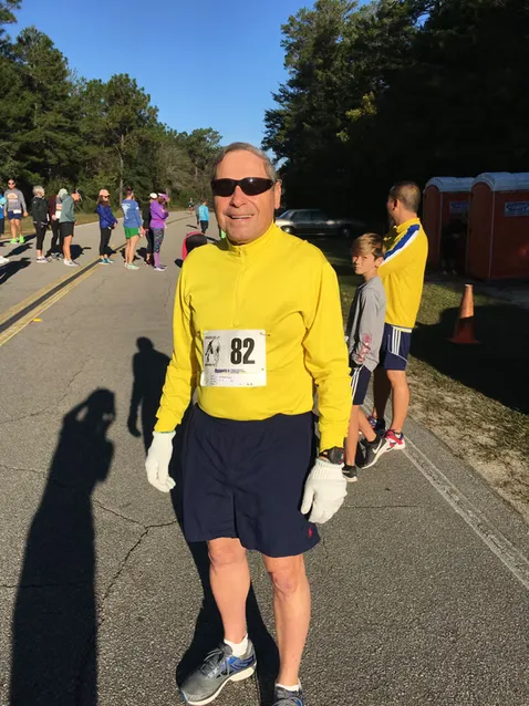 A male runner wearing a yellow long-sleeved shirt, dark shorts, sunglasses, and white gloves, with bib number 82, smiles at the camera. Other runners and portable toilets are visible in the background.