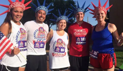 A group of five runners wearing Statue of Liberty crowns and patriotic attire smile for the camera before a race.