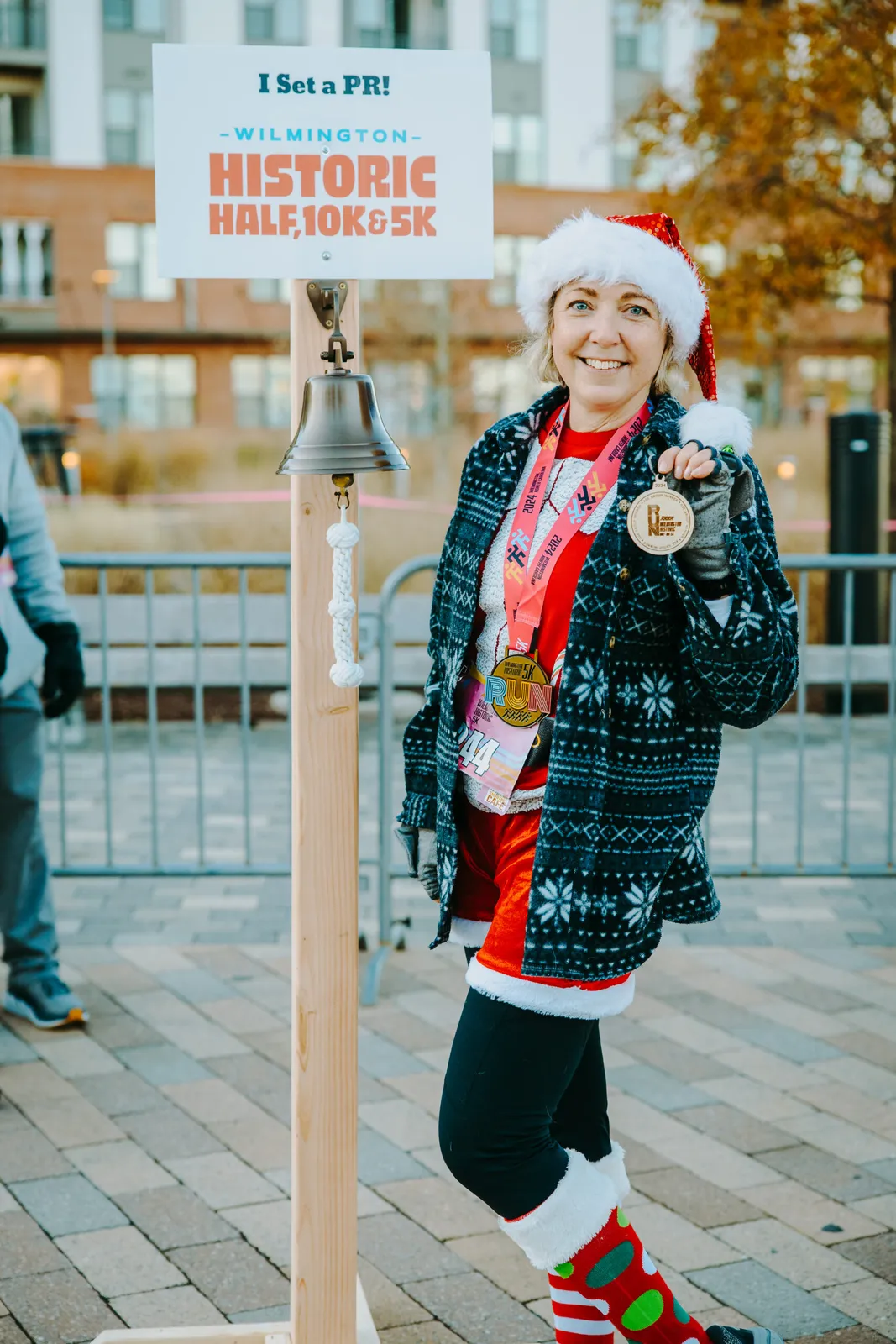 A woman wearing a Santa hat and festive socks smiles as she holds up her race medal and a bell, with a sign behind her that says 'I Set a PR! - Wilmington Historic Half, 10K & 5K'.