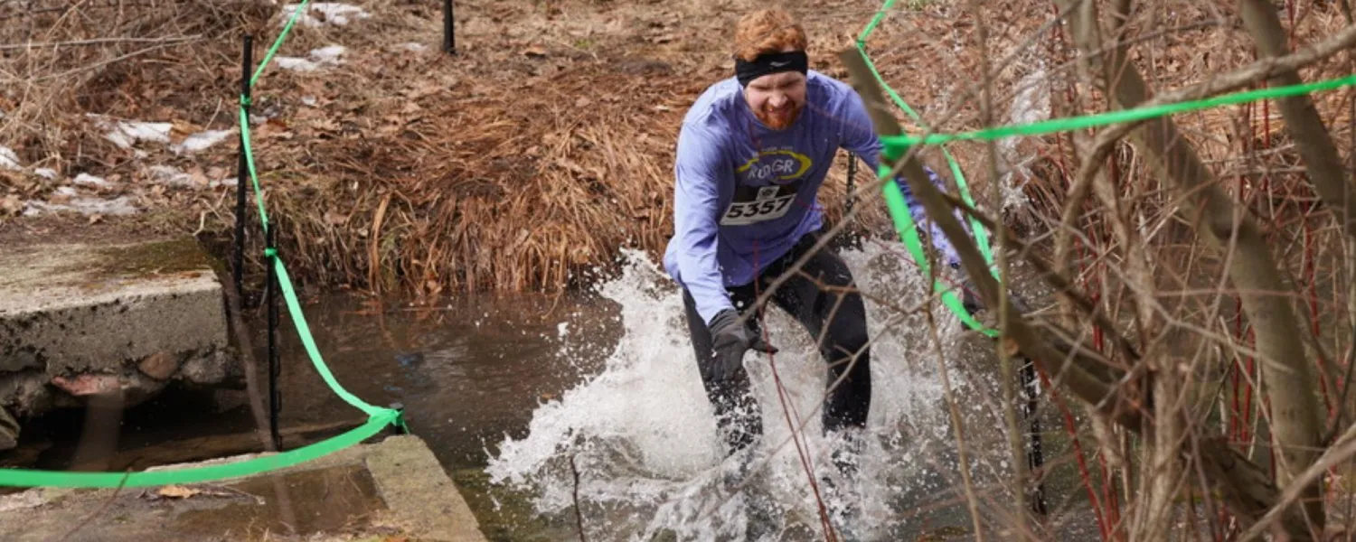 A male runner with a beard and headband splashes through shallow water on a wooded trail, with green marking tape visible.