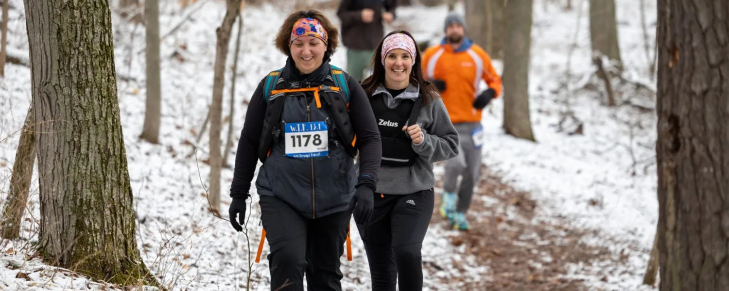 Two women running on a snowy trail, with other runners in the background.