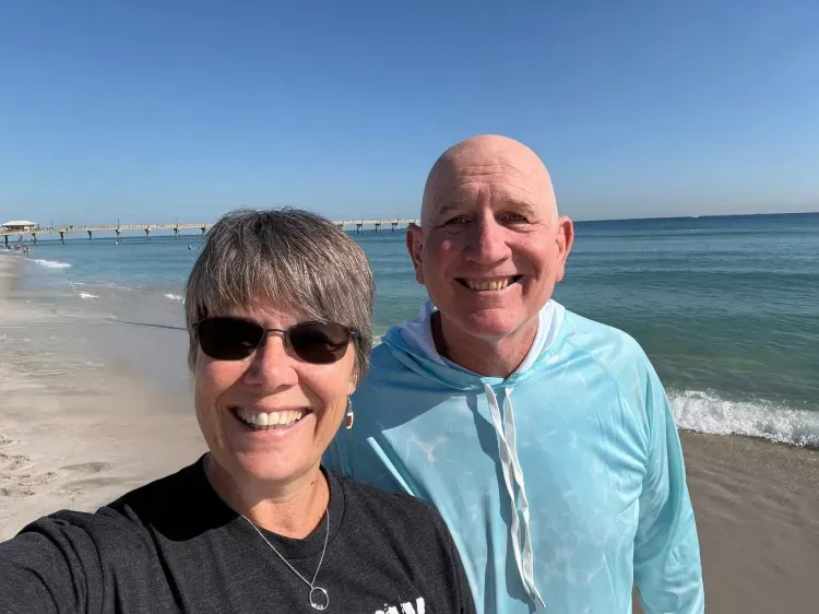 A smiling couple stands on a beach with the ocean and a pier in the background.