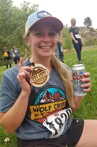 A woman smiles at the camera, holding a wooden medal and a can of Coors Light, with a race bib visible on her shirt.