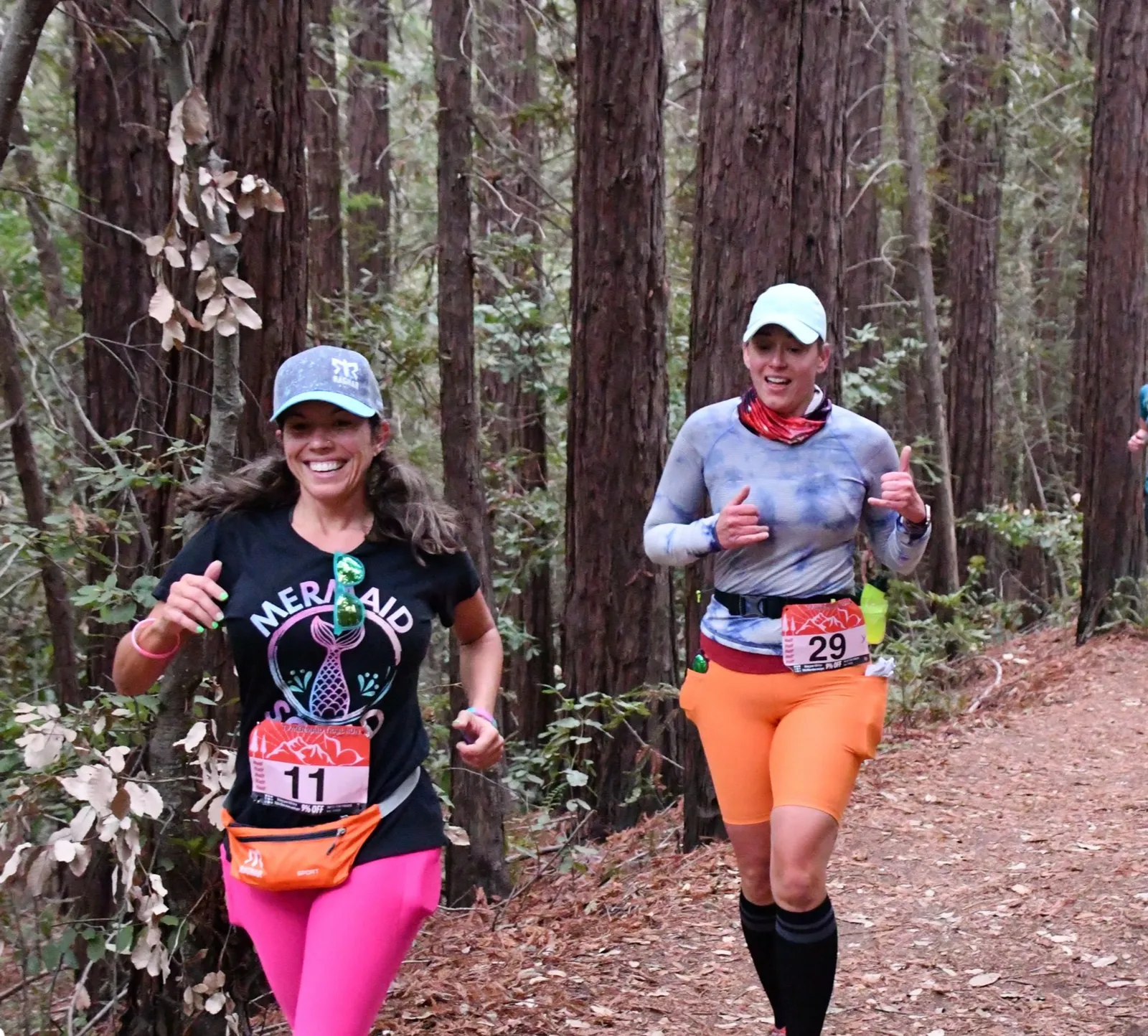 Two women running on a forest trail, both smiling and looking energetic.