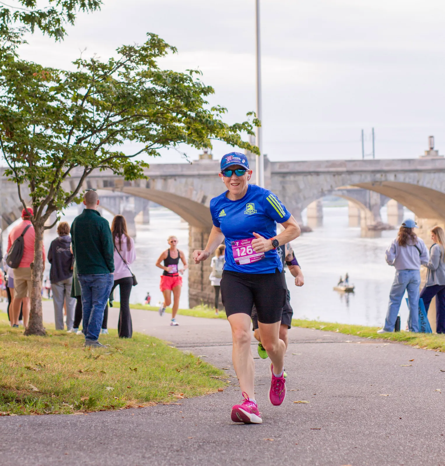 A female runner smiles at the camera while running on a paved path next to a body of water, with a large stone bridge in the background.